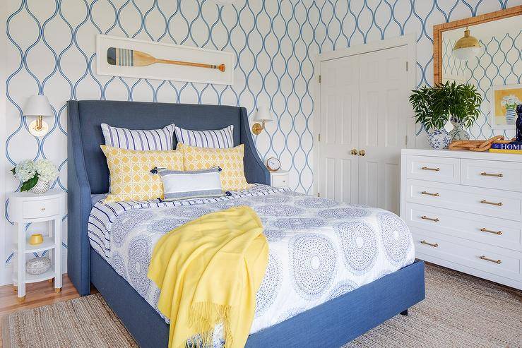 yellow and blue bedroom features an oar art piece hung from a wall covered in blue trellis wallpaper and mounted above a blue wingback bed. The bed, flanked by brass sconces mounted over white oval nightstands, is dressed in blue medallion bedding accented with yellow pillows