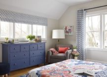 Under a row of windows covered with a single white and blue roman shade, a blue dresser is palced facing a bed dressed in red and blue bedding and placed on a blue rug. A gray rug sits in a corner in front of a black and gold floor lamp lighting light gray bedroom walls.