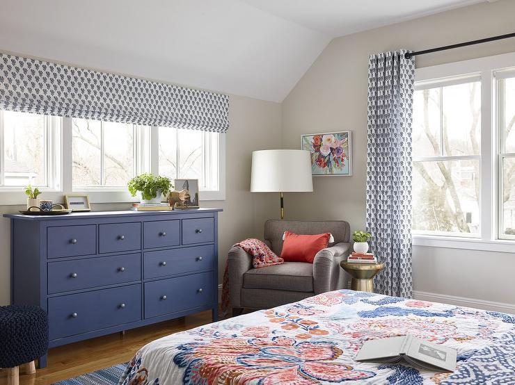 Under a row of windows covered with a single white and blue roman shade, a blue dresser is palced facing a bed dressed in red and blue bedding and placed on a blue rug. A gray rug sits in a corner in front of a black and gold floor lamp lighting light gray bedroom walls.