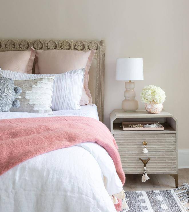 Placed a top a rippled wood nightstand with white tassel pulls, a blush pink lamp lights a carved wood bed. The bed is accented with a pink throw blanket and gray and pink pillows complementing pale pink walls and a gray rug.