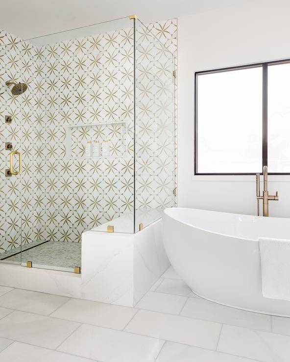 Seamless glass walk-in shower is clad in marble and brass mosaic surround tiles and is finished with a niche. A oval freestanding bathtub is located beside the shower and beneath a window.