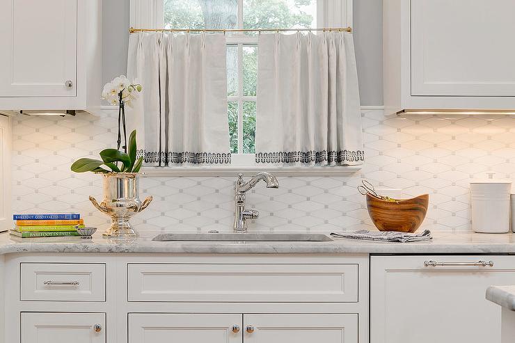 white shaker upper cabinets fixed against white and gray diamond pattern backsplash tiles, a window dressed in white cafe curtains is located over white shaker lower cabinets fitted with a gray quartzite countertop holding a stainless steel sink with a polished nickel faucet.
