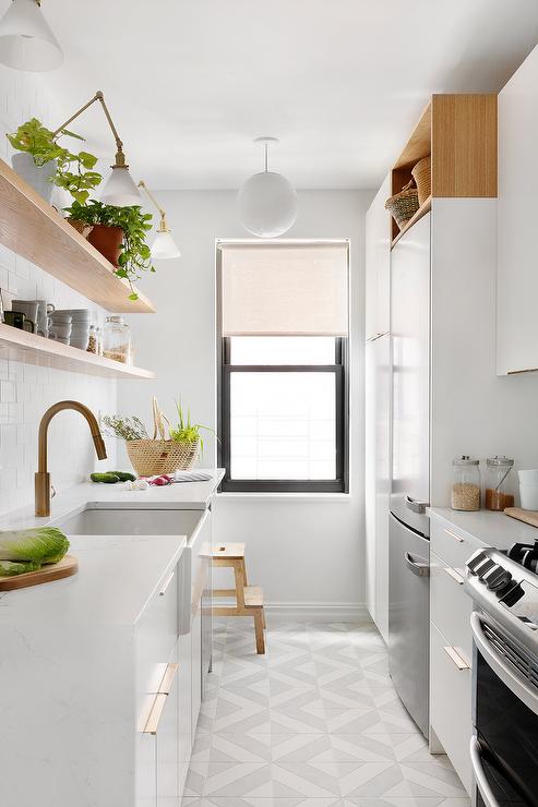 Wood floating shelves lit by brass swing arm sconces are mounted in a galley style kitchen against white subway wall tiles over a farmhouse sink with a brushed gold gooseneck faucet. The faucet is mounted to a marble waterfall countertop accenting white flat front cabinets