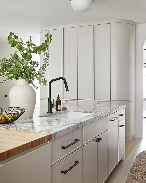 White kitchen island contrasted with matte black pulls features a butcher block and marble countertop holding a sink beneath a matte black faucet.