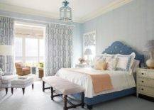 blue French bed accented with blue French bed linens and placed in front of a blue textured wallpapered wall and between cream and blue French nightstands lit by cream gourd lamps. A brown wooden bench sits at the foot of the bed and is lit by a white and blue lantern. Sliding doors opening to a sunroom are covered in white and blue curtains.