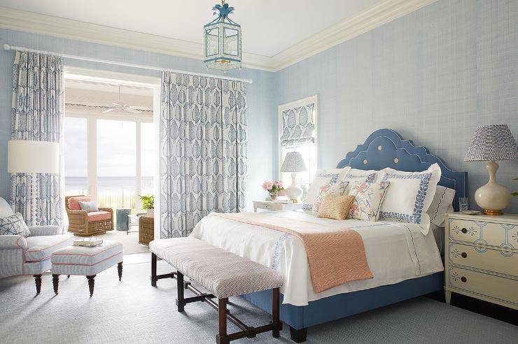 blue French bed accented with blue French bed linens and placed in front of a blue textured wallpapered wall and between cream and blue French nightstands lit by cream gourd lamps. A brown wooden bench sits at the foot of the bed and is lit by a white and blue lantern. Sliding doors opening to a sunroom are covered in white and blue curtains.