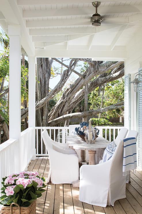 Whitewashed French oval dining table with white slipcovered chairs designed in an outdoor porch in a cottage home