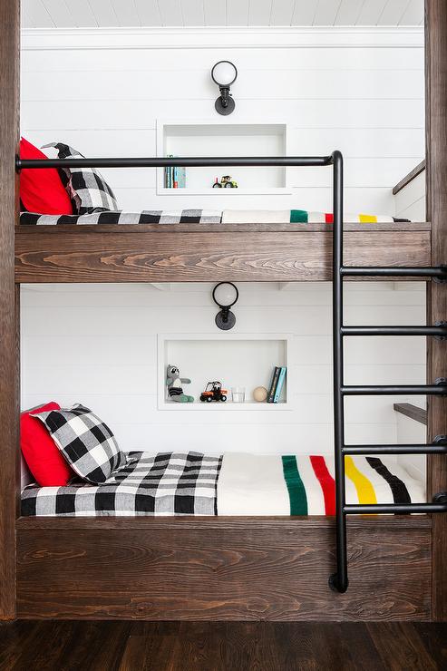 Farmhouse bunk room feature pipe plumbing railing and ladders on dark stained oak wood built-in bunk beds. Black and white plaid bedding mismatch with checkered sheets to create an diverse but stylish look in this shared space. Shiplap trim, wall shelf niches and black sconces complete the styled boys cottage bedroom.