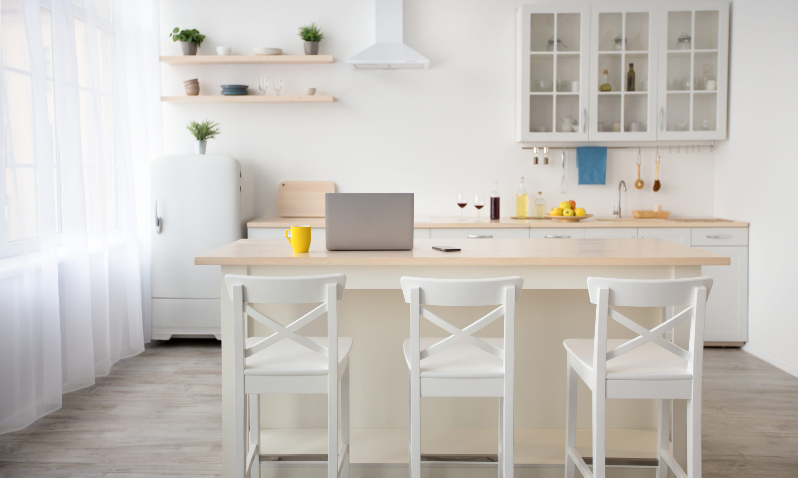 Workspace in kitchen and modern dining room interior. Yellow cup, laptop and smartphone on table, chairs, utensils on furniture, small refrigerator, extractor hood, white walls, curtains in daylight