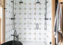 \white and black bath rug in front of a black oval freestanding bathtub matte black tub filler frameless glass shower face to face black vintage style shower heads mounted to white and gray geometric wall tiles over infinity shower drains