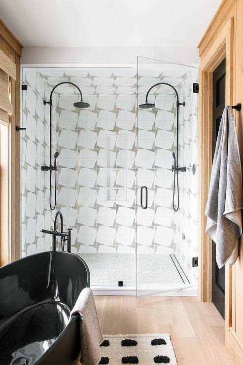 \white and black bath rug in front of a black oval freestanding bathtub matte black tub filler frameless glass shower face to face black vintage style shower heads mounted to white and gray geometric wall tiles over infinity shower drains