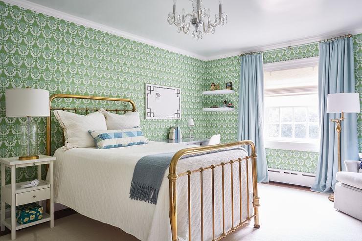 green wallpaper features a brass vintage bed topped with a gray fringe throw blanket and a white and blue lumbar pillow tall white nightstand lit by a glass and brass lamp and a white desk placed beneath a Greek key pin board