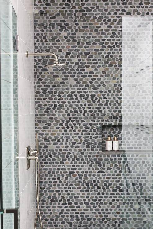 A glass shower door opens to a walk-in shower boasting a stunning gray and black pebble tiled wall framing a pebble tiled niche.
