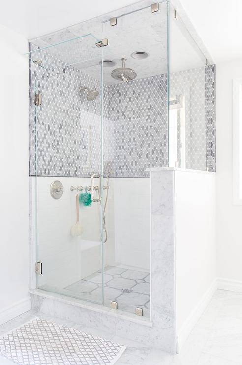 White marble floors topped with a gray trellis bath mat lead to a frameless glass door opening to a walk in marble and glass steam shower fitted with a tilt out window, mosaic marble floors, and gray oval marble tile backsplash holding a polished nickel shower kit and circular shower head while the ceiling holds a rain shower head in this well appointed contemporary bathroom.