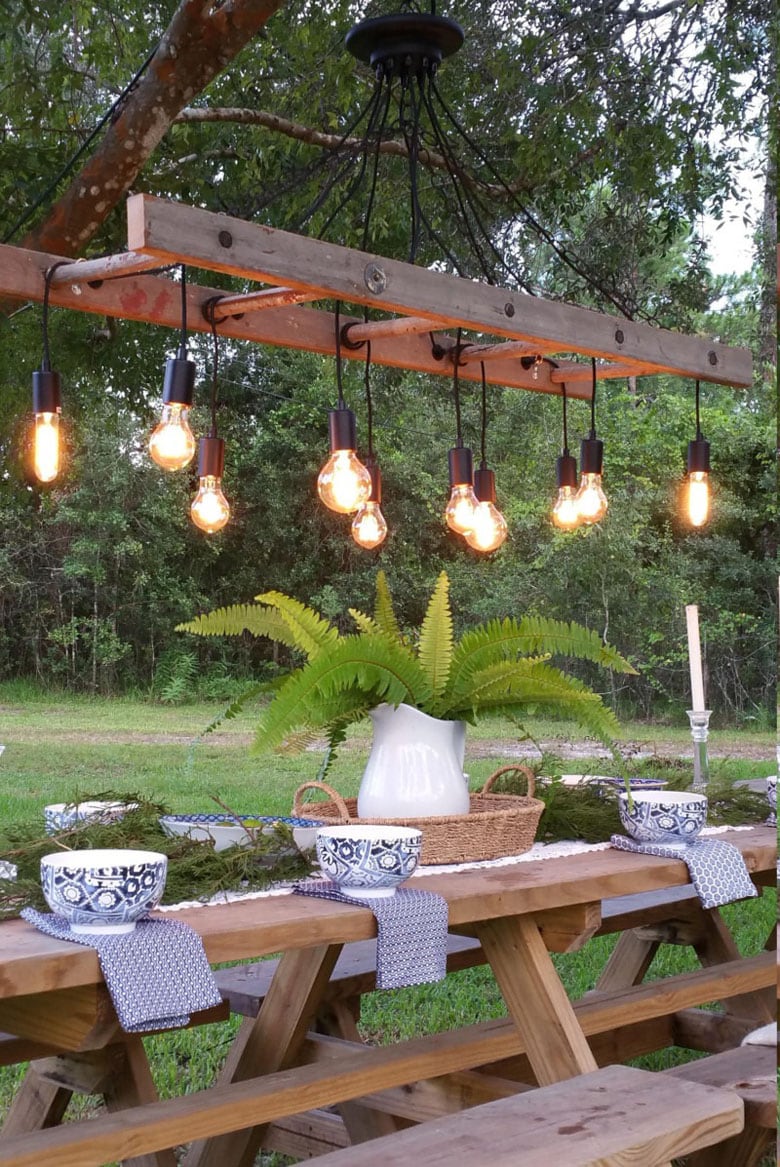 edison bulbs hanging from ladder chandelier outside above dining table with bowls and napkins