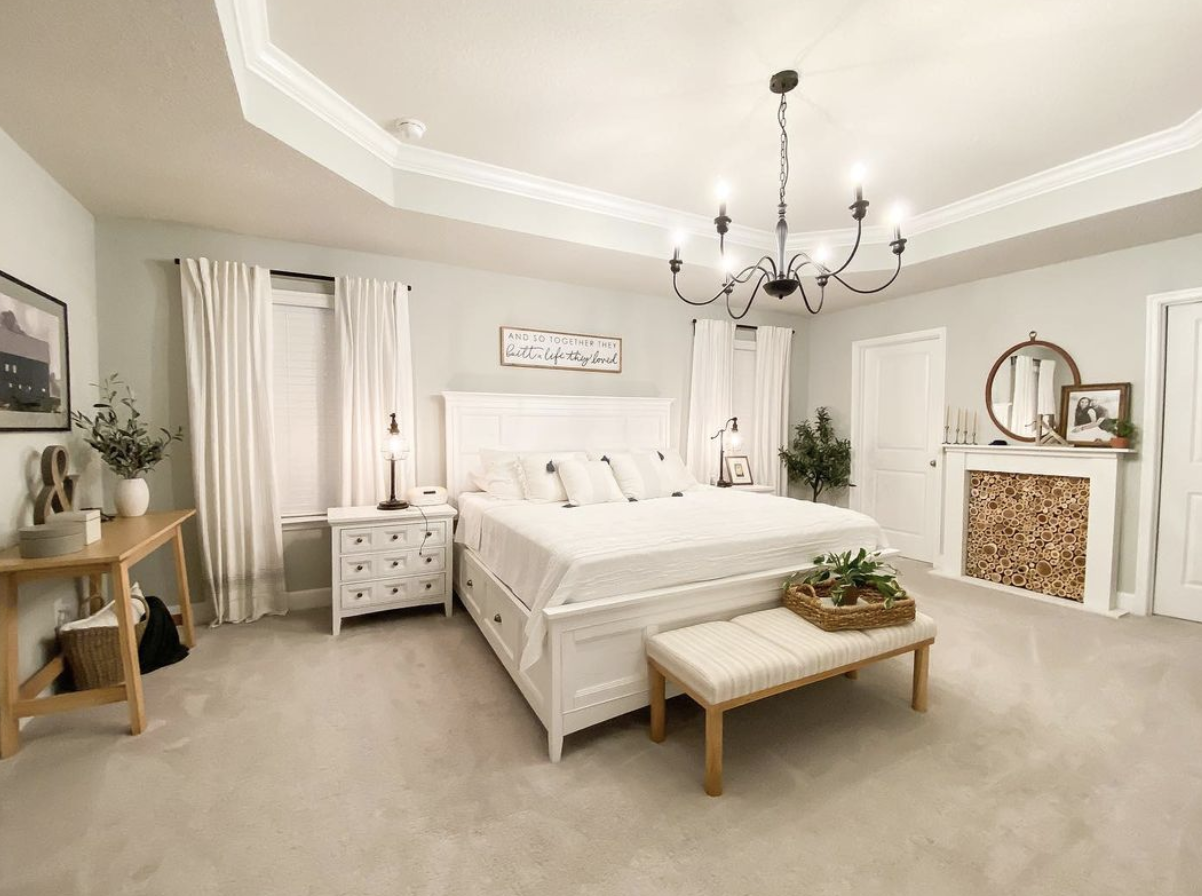 large white master farmhouse bedroom with black chandelier large white bed bench fireplace with wood rounds
