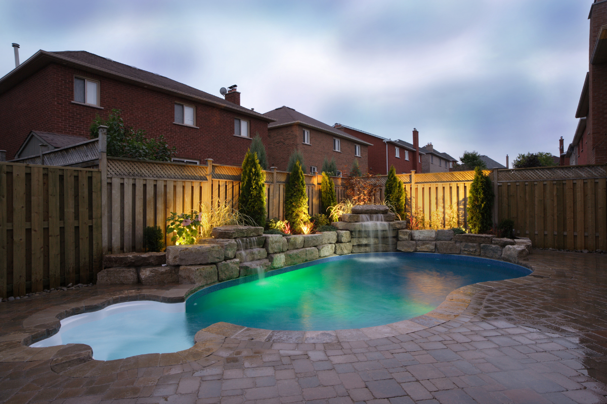 pool backyard lit up stone wall with trees and shrubs houses in background