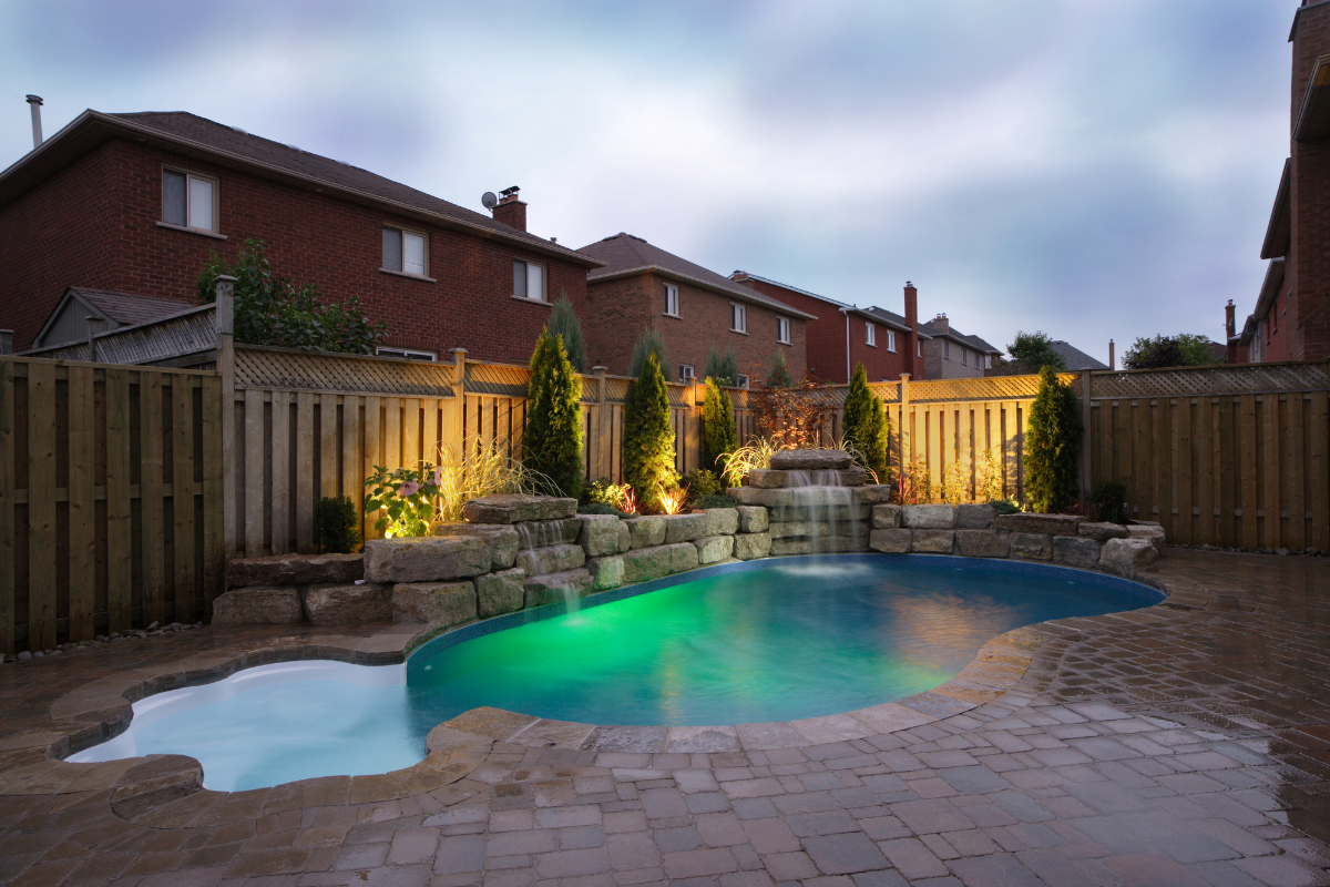 pool backyard with landscape lighting within the stone wall, trees and shrubs. Houses in background.