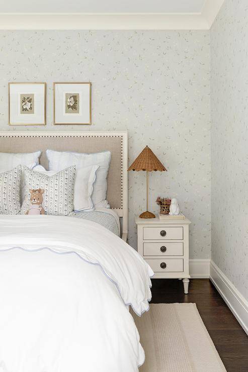 girl's bedroom boasts a white farmhouse nightstand lit by a rattan lamp and placed on a dark stained wood floor beside a bed with a gray burlap headboard. The bed, placed beneath botanical art hung from a pale blue wallpapered wall, is dressed in blue scalloped bedding and sits on a cream rug.