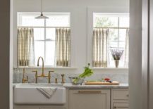 Ivory kitchen features ivory cabinets accented with aged brass hardware and a gray marble countertop, a farm sink with an aged brass deck mount faucet and yellow and gray cafe curtains