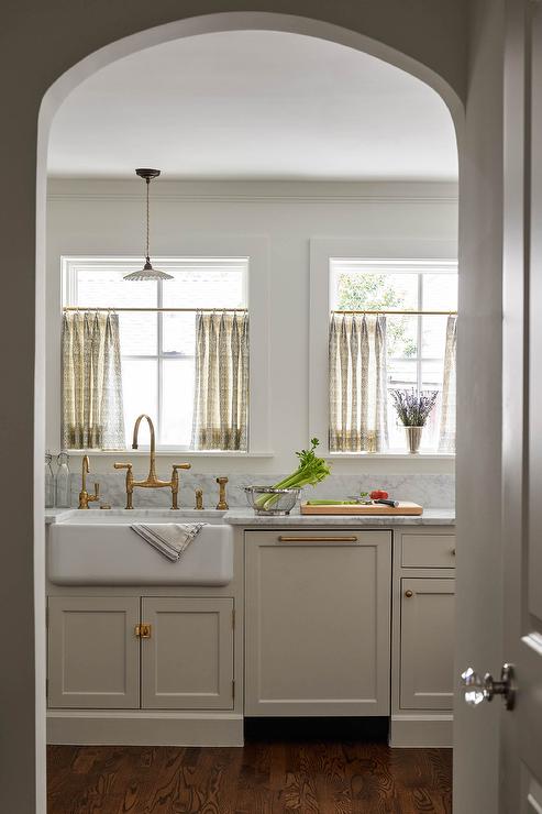 Ivory kitchen features ivory cabinets accented with aged brass hardware and a gray marble countertop, a farm sink with an aged brass deck mount faucet and yellow and gray cafe curtains