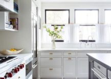Vintage brass cup pulls on white inset cabinets in a transitional kitchen boasting honed white marble countertops and cafe curtains on a brass rod