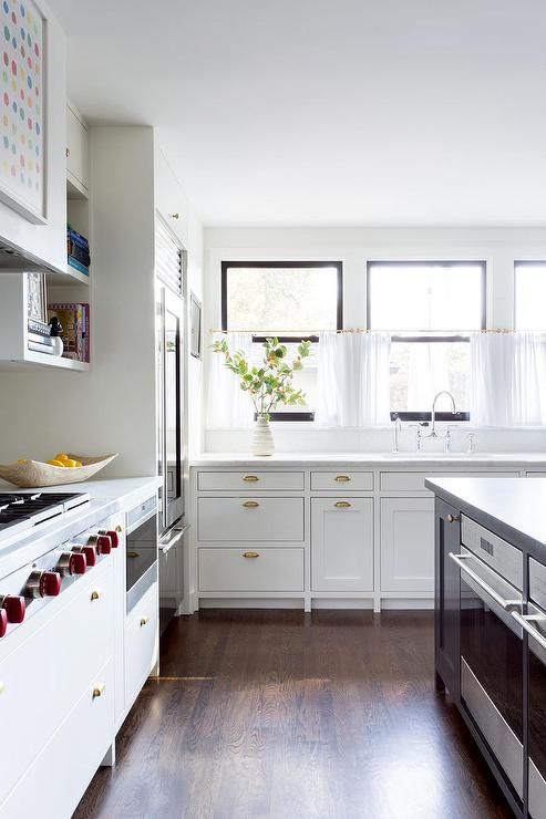 Vintage brass cup pulls on white inset cabinets in a transitional kitchen boasting honed white marble countertops and cafe curtains on a brass rod