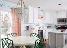 Chandelier hanging over a round off-white dining table paired with green bamboo chairs beside a glass sliding door fitted with orange patterned curtains white cabinetry, a gray island, and white subway backsplash tiles