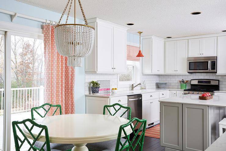 Chandelier hanging over a round off-white dining table paired with green bamboo chairs beside a glass sliding door fitted with orange patterned curtains white cabinetry, a gray island, and white subway backsplash tiles