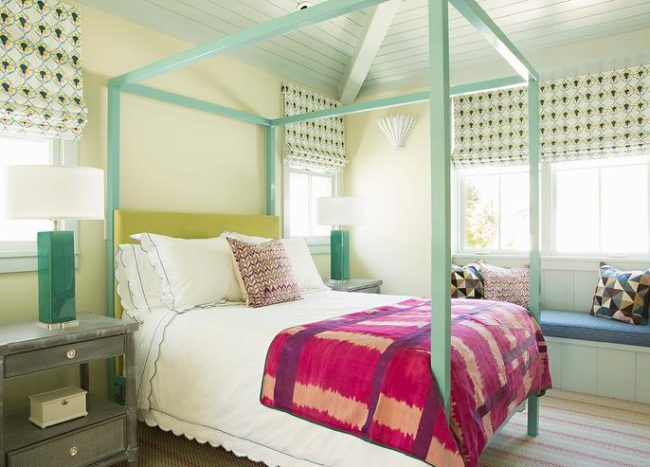calloped bedding topped with a red and purple blanket accents a turquoise blue canopy bed finished with a yellow headboard complementing pale yellow walls. The bed is flanked by green lamps placed on gray nightstands beneath windows dressed in yellow and blue roman shades