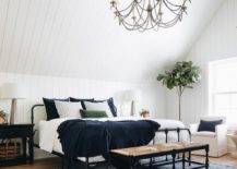 black beaded chandelier hangs from a white plank vaulted ceiling over a black rush seat bench placed on a blue rug at the foot of a black bed dressed in white and dark blue bedding finished with dark blue velvet pillows. In front of a vertical white plank wall, the bed is flanked by black farmhouse nightstand lit by concrete lamps.