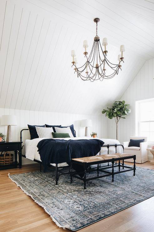 black beaded chandelier hangs from a white plank vaulted ceiling over a black rush seat bench placed on a blue rug at the foot of a black bed dressed in white and dark blue bedding finished with dark blue velvet pillows. In front of a vertical white plank wall, the bed is flanked by black farmhouse nightstand lit by concrete lamps.