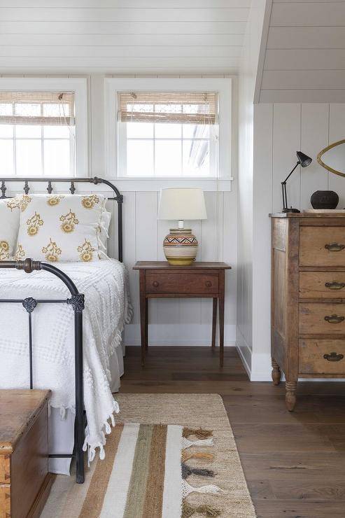 Cottage style bedroom boasting vertical shiplap walls with a row of windows above an oil rubbed bronze bed with white and yellow bedding. A farmhouse style nightstand displays a southwest style lamp complementing layered rugs under the bed.