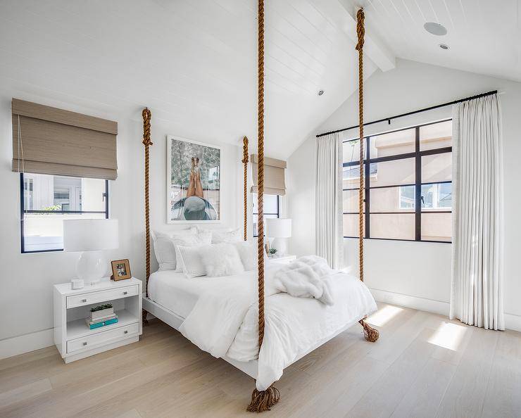 White bedding on a rope hanging bed in a cottage bedroom furnished with white nightstands displaying white lamps. A vaulted white plank ceiling brings a tall appeal to the overall feel of the space with a modern farmhouse finish.