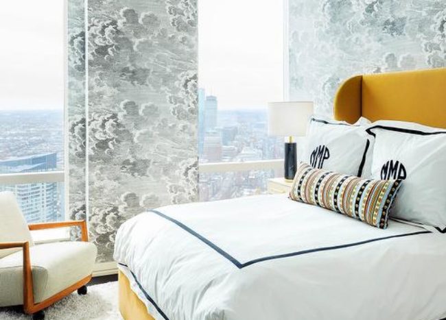 yellow and black contemporary bedroom boasting a mustard yellow velvet bed placed on a gray shag rug and dressed in black border hotel bedding topped with black monogrammed shams. A taupe nightstand lit by black and gold lamp is positioned in front of a wall clad in Fornasetti Nuvole Wallpaper.