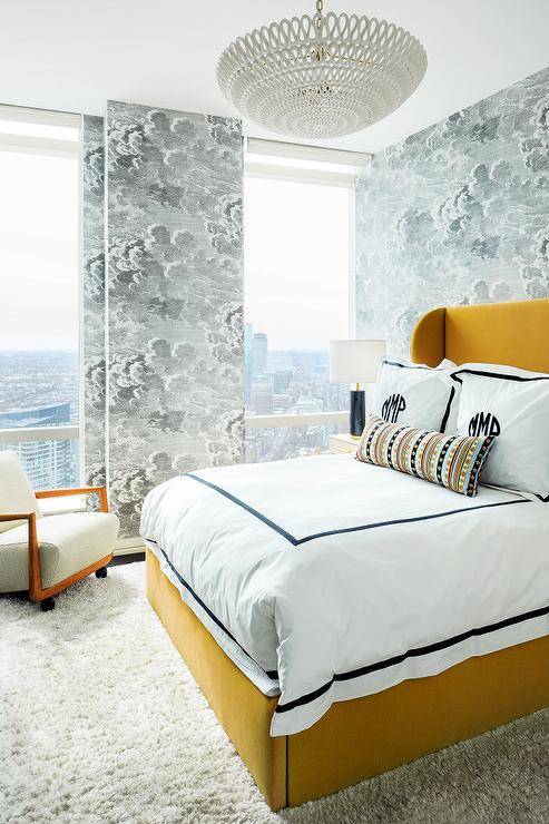 yellow and black contemporary bedroom boasting a mustard yellow velvet bed placed on a gray shag rug and dressed in black border hotel bedding topped with black monogrammed shams. A taupe nightstand lit by black and gold lamp is positioned in front of a wall clad in Fornasetti Nuvole Wallpaper.