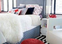 A white sheepskin throw accents the white and red bedding covering a blue geometric bed positioned on a black and white striped rug between windows. Facing a red knitted pouf, a white corner chair is placed beside a white champaign nightstand lit with a silver lamp.