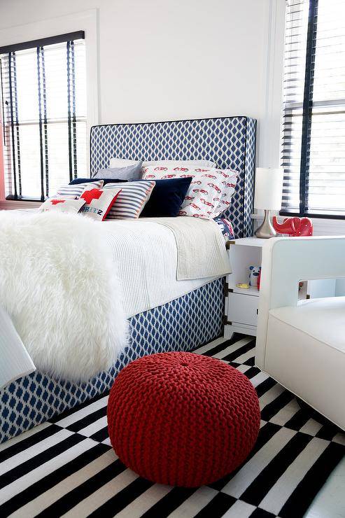 A white sheepskin throw accents the white and red bedding covering a blue geometric bed positioned on a black and white striped rug between windows. Facing a red knitted pouf, a white corner chair is placed beside a white champaign nightstand lit with a silver lamp.