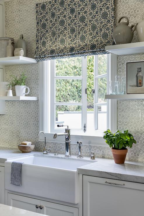 pattern roman shades hanging over white kitchen window pattern wallpaper white apron farmhouse sink open shelving potted plant on counter