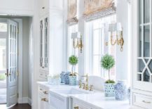 White Chippendale kitchen cabinets are adorned with antique brass pulls and hold a farmhouse sink beneath a polished nicke hook and spout faucet fixed to a white marble countertop beneath a window dressed in beige curtains and flanked by brass 2-light sconces. Mullion cabinets are located on each end of the wall.