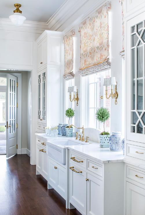 White Chippendale kitchen cabinets are adorned with antique brass pulls and hold a farmhouse sink beneath a polished nicke hook and spout faucet fixed to a white marble countertop beneath a window dressed in beige curtains and flanked by brass 2-light sconces. Mullion cabinets are located on each end of the wall.