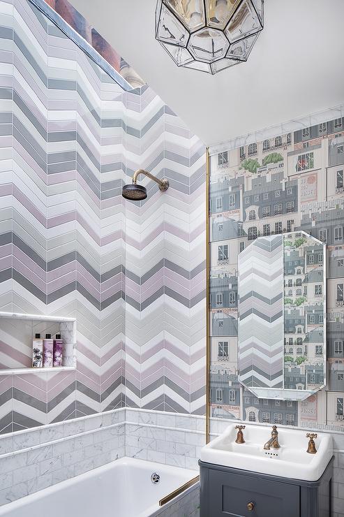 Bathroom features a shower with pink and gray chevron pattern tiles, a marble subway tiled drop-in bathtub, and a beveled mirror on gray French house print wallpaper over a gray single washstand with antique brass vintage cross handle faucet