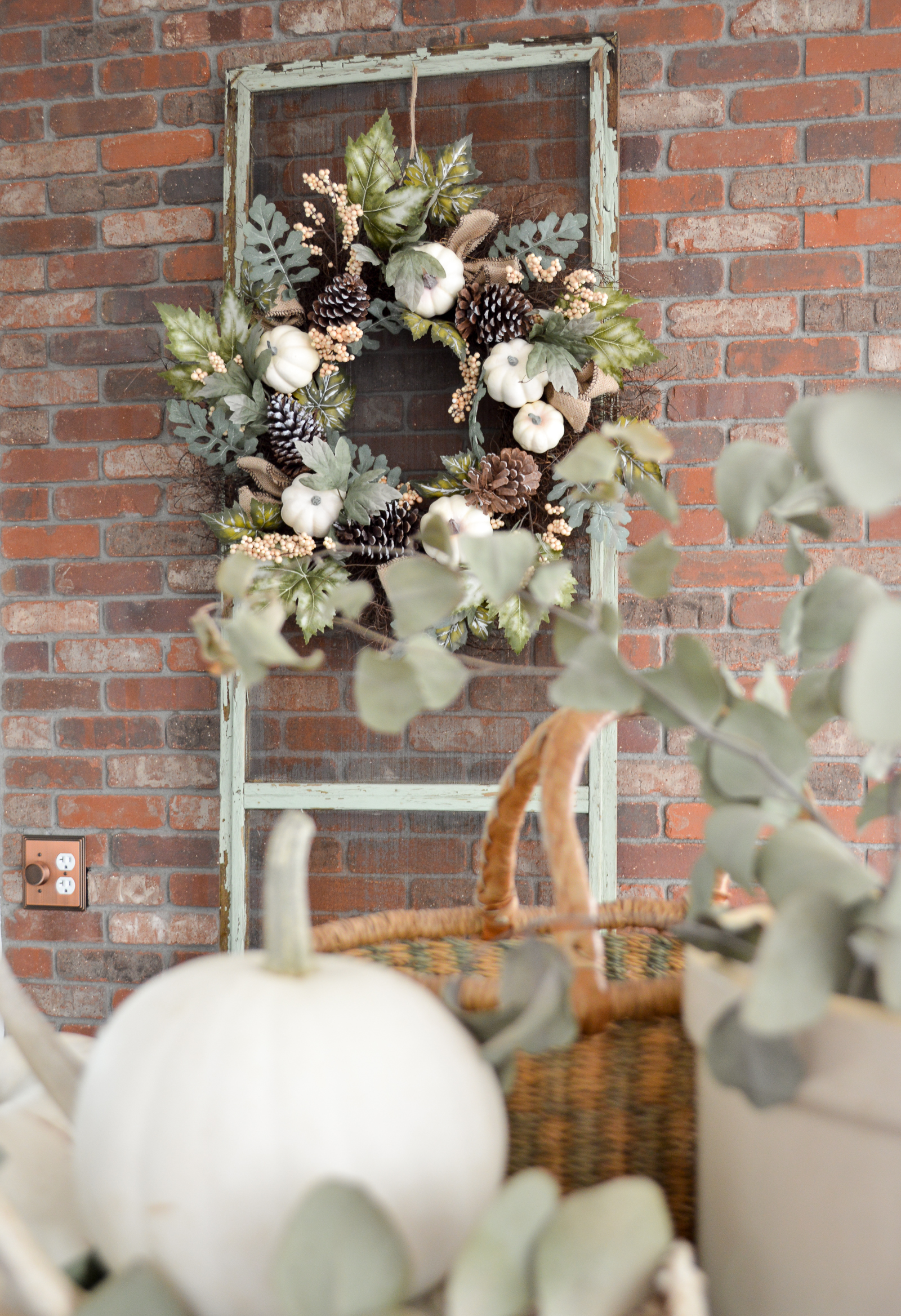 fall wreath hanging on old screen window up against red brick with pumpkin in foreground