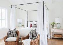 cottage bedroom features a vaulted ceiling with white wood beams over an iron canopy bed fitted with a tan headboard accented with white sheers flanked by light wood farmhouse nightstands topped with recycled glass lamps alongside a pair of seagrass chairs accented with black and white pillows placed at the foot of the bed atop a jute rug.