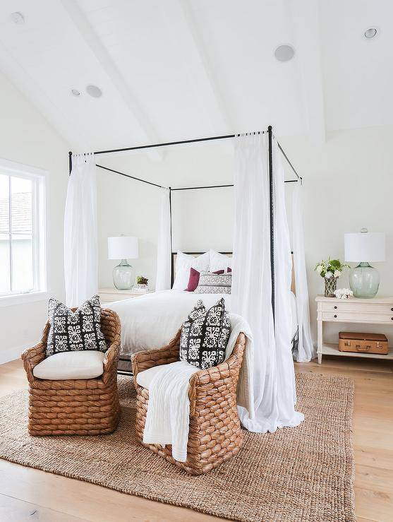 cottage bedroom features a vaulted ceiling with white wood beams over an iron canopy bed fitted with a tan headboard accented with white sheers flanked by light wood farmhouse nightstands topped with recycled glass lamps alongside a pair of seagrass chairs accented with black and white pillows placed at the foot of the bed atop a jute rug.