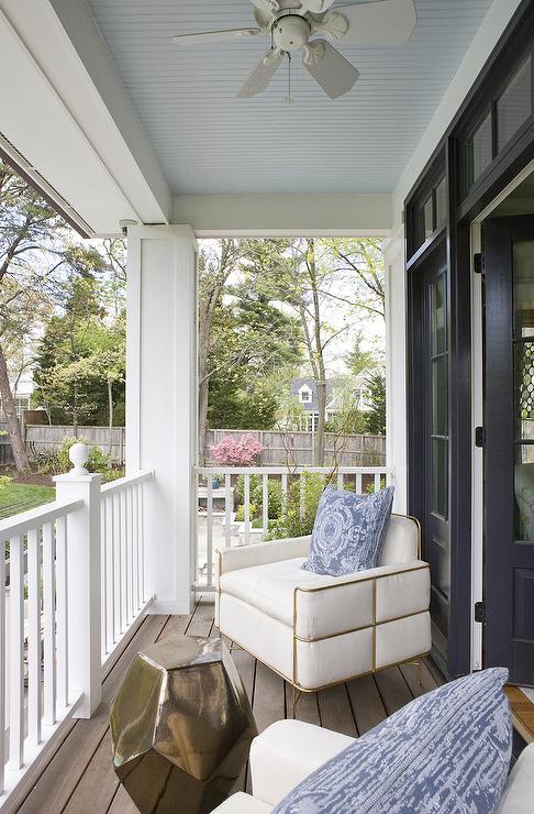 Gorgeous covered porch features a blue beadboard ceiling accented with a ceiling fan over ivory upholstered chairs with brass trim adorned with blue medallion pillows alongside a brass geometric stool table.