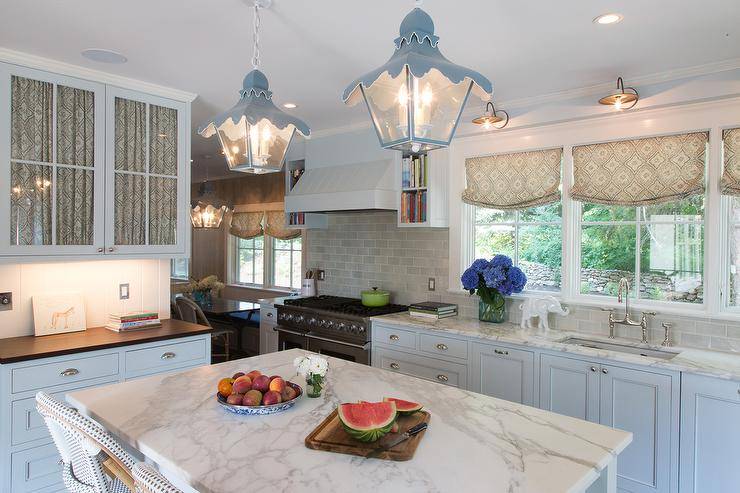 Kitchen with white cabinets paired with statuary marble counters and a gray glazed subway tiled backsplash. A white paneled kitchen hood, flanked by open cookbook shelves, stands over a stainless steel stove. A stainless steel kitchen sink sits under three windows illuminated by bronze sconces.