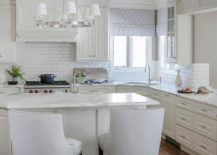 ivory kitchen with an angled island topped with a white and gray marble countertop seats white French stools facing an island sink. Light gray trellis curtains hang over a corner sink paired with a polished nickel gooseneck faucet.