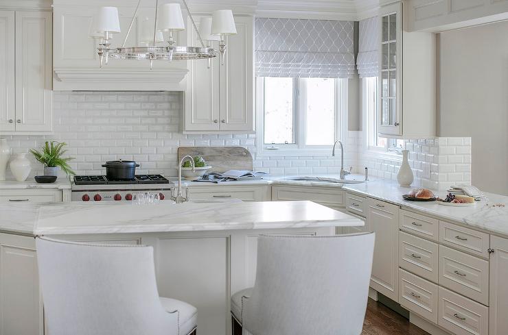 ivory kitchen with an angled island topped with a white and gray marble countertop seats white French stools facing an island sink. Light gray trellis curtains hang over a corner sink paired with a polished nickel gooseneck faucet.