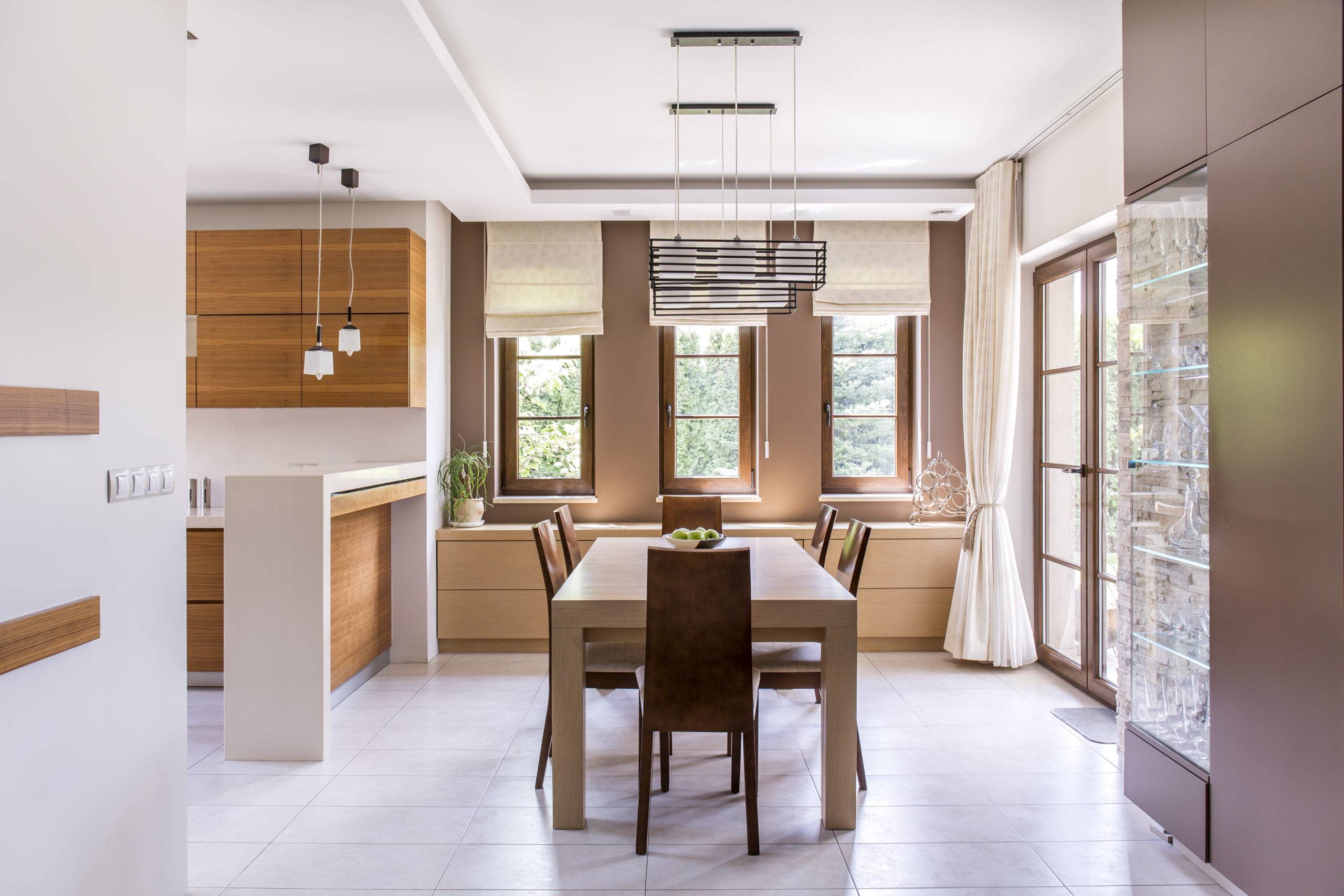 Kitchen and dining room in modern house with wood table and chairs three long skinny windows wood cupboards and sleek design contemporary chandelier